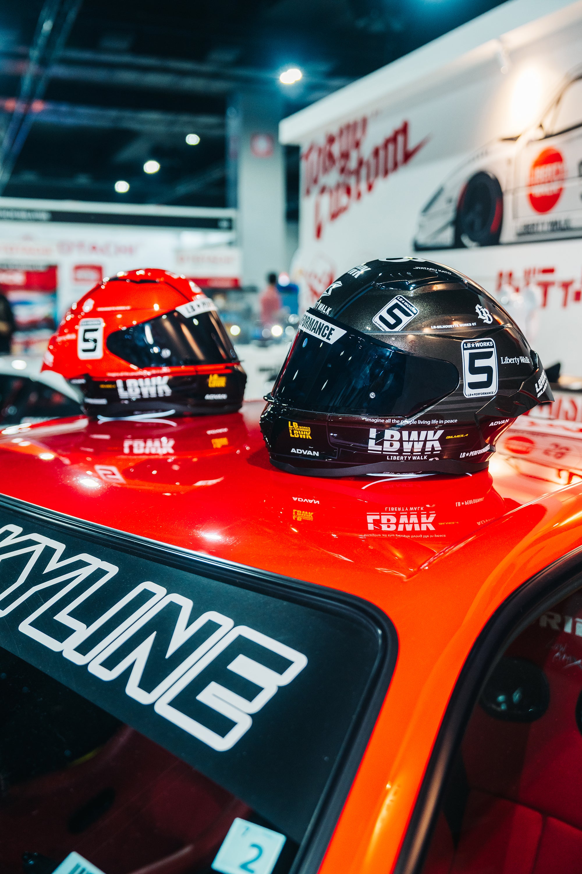 Two R34 Bilmola motorcycle helmets, one red and one black, displayed on the roof of the Liberty Walk R34 race car at an exhibition show. The helmets feature the number 5 and branding for LBWK.